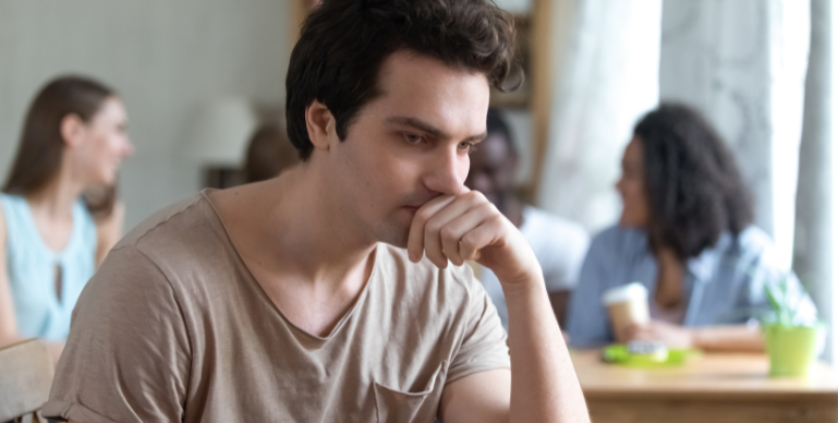 Young white male looking reflective with his hand to his face while sitting at a table with people in the background
