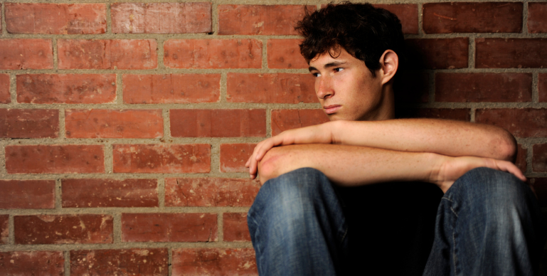 male teen sitting against a brick wall looking sad and contemplative