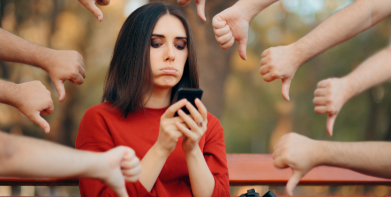 white woman sits on a bench looking at her phone with her bottom lip curled down. In the background around her several arms/hands are extended with a thumbs down gesture