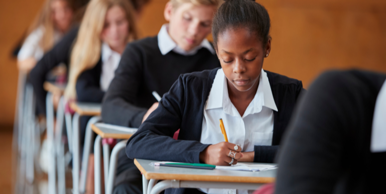 a black teenager sits at her desk in a classroom, writing on paper with a pencil. Students similarly dressed in a private school uniform can be seen behind her in the background.