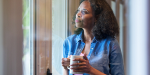 woman holds a mug with both hands and gazes outside in front of a window with a slight smile on her face