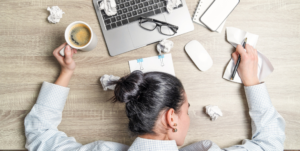 woman with dark hair in a bun has her face down on a messy desk with a laptop, glasses, papers strewn about