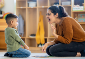 a women sits on the ground of a playroom facing child pointing to her smile; teaching children emotions and being aware of feelings
