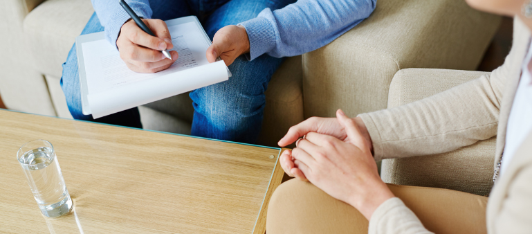 two people are sitting together and only their hands are visible, one holding a clipboard, the other has hands clasped together on their lap (waiting too long to get an adhd assessment)