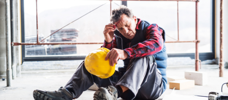 (ptsd can coincide with chronic pain) a worker sits on the ground holding his helmet with one hand and with the other touches his head while closing his eyes tight