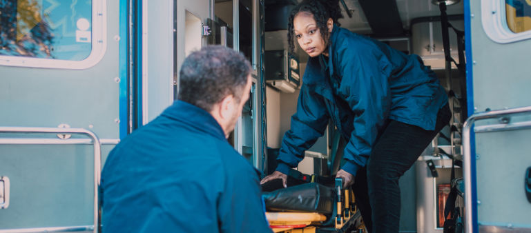 a women paramedic helps unload a stretcher from the back of an ambulance with a male paramedic on the ground (paramedics high risk of trauma)