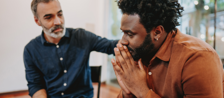 a man appears thoughtful as he puts his hands together in front of his face, a man sitting beside him looks to him with a hand on his shoulder (peer support for first responders)